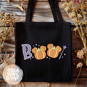 May include: Black tote bag with the word "Boo" embroidered in purple and orange. The "OOs" are designed as Mickey Mouse pumpkins, with a purple wand and stars. The bag has two black handles and a "Stitch Love Craft" logo.