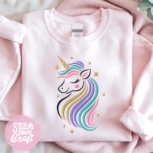 Puede incluir: Una sudadera rosa claro con un diseño de unicornio colorido. El unicornio tiene un cuerno dorado, una melena en tonos azules, rosas, morados y dorados, y un rubor en forma de corazón. Estrellas doradas rodean el unicornio. El texto "Stitch Love Craft" está en un círculo rosa.