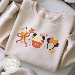 Op de afbeelding: Een crèmekleurige sweater met ronde hals en een geborduurd ontwerp van een koffiebeker met een rode strik, een cupcake met muizenoortjes en een haarband met muizenoortjes, geaccentueerd met kleine oranje herfstblaadjes en rode hartjes.