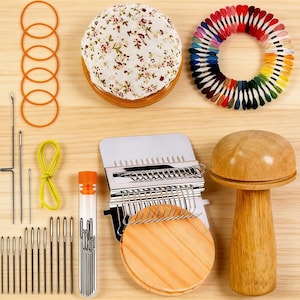 May include: A collection of sewing and embroidery supplies, including a floral pin cushion, a ring of colorful embroidery floss, needles, rubber bands, and a wooden mushroom-shaped tool. The items are arranged on a light wood surface.