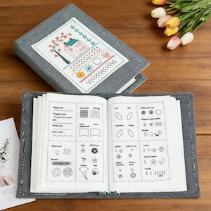 May include: A gray fabric-covered book with an embroidered cover featuring a house and tree design. The open book displays various embroidery stitch examples in black ink. The book is on a wooden surface.