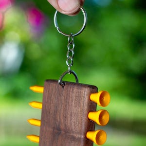 May include: A rectangular dark brown wooden keyring featuring five bright yellow golf tees inserted into each side. The wooden block is attached to a silver metal split ring and chain, creating a functional accessory for golf enthusiasts.