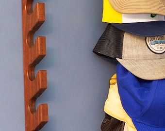 Puede incluir: Un perchero vertical de madera montado en la pared con múltiples muescas para colgar objetos, con un acabado marrón oscuro. Una pila de cinco gorras de béisbol de varios colores y diseños, incluida una con un parche de Tatooine, cuelga del perchero.