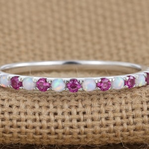 May include: A silver ring with a band of alternating pink and white gemstones. The gemstones are round and evenly spaced around the ring. The ring is set against a textured, tan background.
