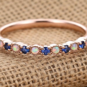 May include: Rose gold ring with alternating opal and sapphire gemstones. The ring features a delicate band and a row of small, round gemstones in white, blue, and iridescent colors. The ring is set against a neutral, textured background.