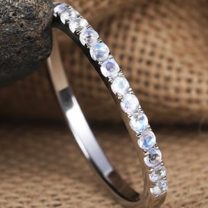 Könnte beinhalten: Ein dünner silberfarbener Ring mit einer Reihe kleiner, runder, durchscheinender Mondsteine, die einen dezenten blauen Schimmer aufweisen. Die Steine sind in einer durchgehenden Linie entlang der oberen Hälfte des Metallrings gefasst und wirken fein.