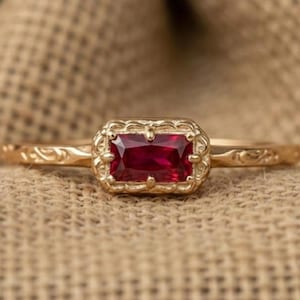 Könnte beinhalten: Ein goldener Ring mit einem rechteckigen, tiefroten Edelstein. Der Ring hat eine verzierte Fassung im Vintage-Stil und ein zartes, graviertes Band. Der Edelstein ist der Mittelpunkt, eingebettet in einen detaillierten Rahmen.
