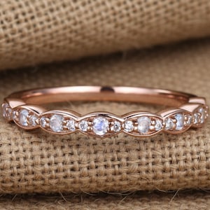 Könnte beinhalten: Ein Ring aus Roségold mit einer Reihe abwechselnder runder Mondsteine und kleinerer klarer Akzentsteine in einem gewellten Metalldesign. Der Ring hat eine polierte Oberfläche und ein zartes, schmales Profil mit einer durchgehenden Edelsteinreihe.