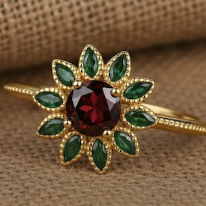 May include: A gold ring featuring a central round dark red garnet stone surrounded by a halo of twelve marquise-cut green stones arranged in a floral pattern. The band has a delicate beaded texture along the outer edges of the metal setting.