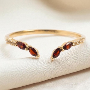 Könnte beinhalten: Ein goldfarbener offener Ring mit drei ovalen, tiefroten Edelsteinen auf jeder Seite. Der Ring hat ein zartes Band und kleine, klare Akzentsteine in der Nähe der roten Edelsteine. Der Ring ist vor einem weichen, cremefarbenen Hintergrund platziert.