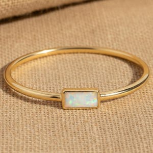 May include: A gold bangle bracelet featuring a rectangular opal stone. The opal has a white, iridescent appearance. The bracelet is displayed on a tan surface.