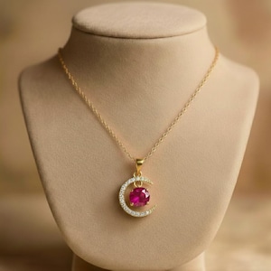 Könnte beinhalten: Eine goldene Halskette mit einem halbmondförmigen Anhänger. Der Mond ist mit kleinen klaren Steinen besetzt und hält in der Mitte einen runden, leuchtend fuchsia-pinkfarbenen Edelstein. Das Metall hat durchgehend ein poliertes Gold-Finish.