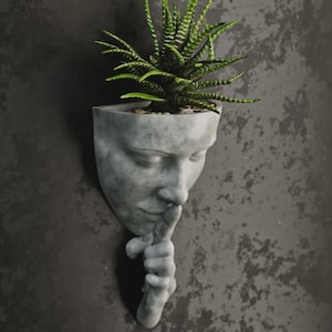 Könnte beinhalten: Ein grauer Wandtopf aus Beton in Form eines menschlichen Gesichts, mit einem Finger auf den Lippen in einer „Psst“-Geste. Der Pflanztopf enthält eine grüne Sukkulentenpflanze. Die Skulptur ist an einer strukturierten grauen Wand befestigt.