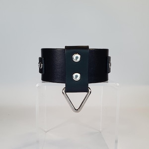 Può includere: Un bracciale da polso in pelle nera con un anello triangolare in metallo color argento. Il bracciale è fissato con due rivetti color argento e una chiusura a fibbia. Il bracciale è largo circa 3,8 cm.