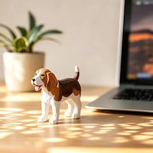 Op de afbeelding: Een houten beagle beeldje, met de hand gesneden en beschilderd met bruine, witte en bruine details. De hond staat op een houten oppervlak, met een laptop en een plant in een pot op de achtergrond. Het beeldje is ongeveer 13 cm lang.