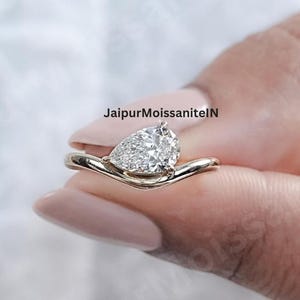 May include: A gold ring with a pear-shaped, clear gemstone. The ring has a unique, curved band design. The text "JaipurMoissaniteN" is visible in the background. The ring is held up by a person's fingers.
