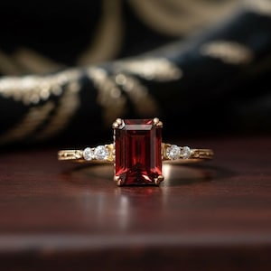 May include: A gold ring with a rectangular, deep red gemstone as the focal point. The ring is accented with smaller, round, clear gemstones on each side of the main stone. The band has a delicate, patterned design.