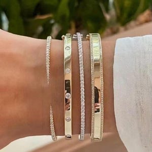 Può includere: Una collezione di braccialetti d'oro con dettagli di diamanti. I braccialetti variano nello stile, tra cui una sottile fascia tempestata di diamanti, una fascia d'oro con diamanti e una fascia d'oro più larga. I braccialetti sono indossati su un polso.