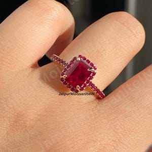 Könnte beinhalten: Ein goldener Ring mit einem großen, rechteckigen, roten Edelstein, umgeben von kleineren roten Steinen. Der Ring hat auch kleine rote Steine am Band. Der Ring befindet sich an einem Finger.