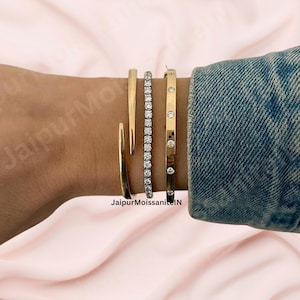 Könnte beinhalten: Eine Auswahl an goldenen Armbändern und ein Diamant-Tennisarmband werden an einem Handgelenk gezeigt. Die Armbänder umfassen einen offenen Armreif, ein diamantbesetztes Band und ein Scharnierarmband mit runden Details. Ein Jeansjackenärmel bedeckt das Handgelenk teilweise.