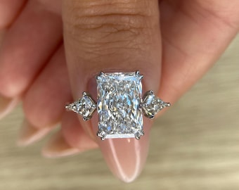 May include: A radiant-cut diamond ring with two smaller, triangular diamonds on either side. The large, rectangular center stone sparkles with numerous facets, set in a silver or white gold band. The ring is held up against a neutral background.