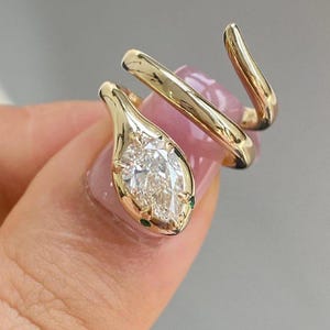 Puede incluir: Anillo de serpiente dorado con una piedra preciosa clara en forma de pera como cabeza de serpiente. El anillo se enrolla alrededor del dedo con un diseño curvo. Pequeñas piedras verdes acentúan la cabeza de la serpiente.