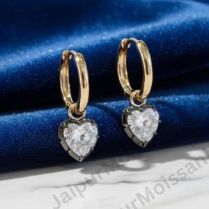 May include: A pair of gold hoop earrings with heart-shaped pendants. The heart-shaped pendants are white with a dark grey border. The earrings are displayed on a blue velvet surface.
