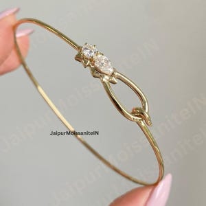 Könnte beinhalten: Ein zartes goldenes Armreif-Armband mit zwei klaren, funkelnden Edelsteinen. Das Armband hat ein einzigartiges Design mit einem knotenartigen Verschluss. Es wird von einer Person gehalten.