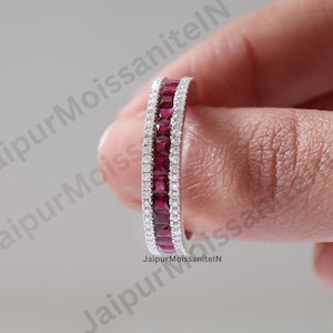 May include: A silver ring featuring a row of square, deep red gemstones and a border of small, clear stones. The ring is held between fingers, highlighting the detailed design and sparkle.
