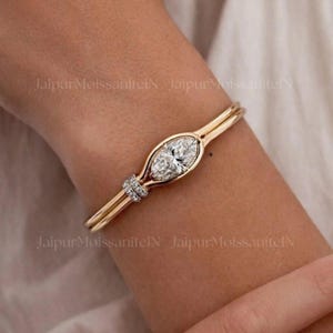 Puede incluir: Un brazalete rígido de oro con una piedra central de talla marquesa en engaste de bisel, flanqueada por pequeñas piedras en pavé en un lado. La banda de metal es lisa y pulida, creando un diseño de bucle continuo sobre la piel.
