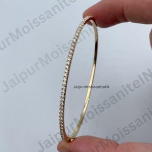 Könnte beinhalten: Ein goldener Armreif mit einer Reihe kleiner, klarer Edelsteine. Das Armband wird von einer Hand gehalten. Der Text "JaipurMoissaniteIN" ist auf dem Armband zu sehen.