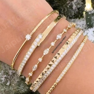May include: A collection of gold bracelets, each with a unique design. Some are adorned with clear gemstones, while others feature sleek, minimalist bands. The bracelets are displayed on a wrist, showcasing their elegant appeal.