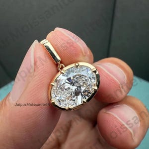 Puede incluir: Un colgante de oro con una gran piedra preciosa clara de talla ovalada. La piedra preciosa está engastada en un bisel de oro con un pequeño lazo para una cadena. El colgante está en una mano. El texto "JaipurMoissaniteIN" es visible.