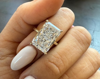 Op de afbeelding: Een gouden ring met een rechthoekige diamant. De diamant schittert door zijn facetten. De ring wordt getoond op een hand met witte nagellak, wat een contrast creëert. Een opvallend sieraad.