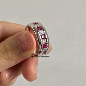 May include: A silver ring with alternating square-cut red and white gemstones. The ring is held in a hand, showcasing the polished metal and the vibrant colors of the stones. The ring is a piece of jewelry.