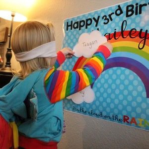May include: A child with a blindfold is playing a party game, trying to pin a cloud onto a rainbow poster. The poster says "Happy 3rd Birthday Bailey" and "Pin the cloud on the Rainbow". The child's sleeves are rainbow-striped.