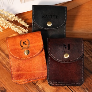 May include: Three leather cases in black, brown, and orange, each with a flap closure and a brass snap. The cases are personalised with names and initials. The orange case has the name "Kathie" and the initial "K" inside a wreath.