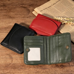 May include: Three leather wallets in black, red, and green are displayed on a wooden surface. The green wallet is open, revealing card slots and a clear ID window. The red and black wallets are closed, showcasing their curved flap design. A newspaper is in the background.