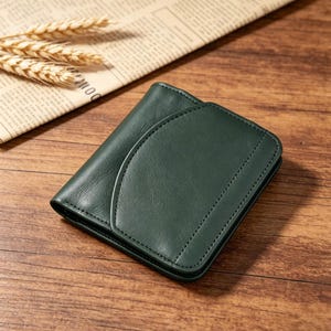 May include: A dark green leather wallet with a curved flap closure. The wallet is rectangular and features stitched detailing along the edges. The background includes a newspaper and wheat stalks on a wooden surface.