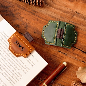 Può includere: Due segnalibri in pelle con cuciture decorative e fasce elastiche. Uno è marrone con le iniziali "M♥S" e l'altro è verde. I segnalibri sono su una superficie di legno con una penna e un libro.