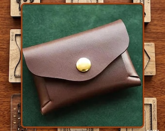 May include: A brown leather cardholder with a curved flap and a gold button closure. The cardholder is set against a green background, with wooden craft tools surrounding it. The leather appears smooth and well-crafted.