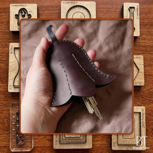 May include: A dark purple leather key holder in a tulip shape, with white stitching and a small loop. The key holder contains several keys. The background includes wooden craft tools and the "Brandy Time" logo.