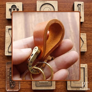 May include: A tan leather keychain with a gold-colored clasp and two gold rings. The keychain is held in a hand, with several wooden craft tools in the background. The clasp has the word "FOX" and "EING" engraved on it.