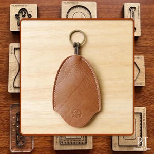 May include: A brown leather key holder with a stitched border and a metal ring. The key holder has a rounded bottom and a small embossed design. It is displayed on a wooden surface with various wooden craft tools.