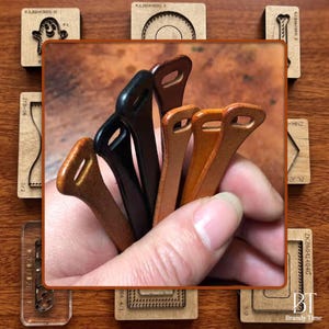 May include: A collection of leather craft tools in various shades of brown and black. The tools have a loop at the top and are held in a hand. Wooden stamps with various designs surround the tools.