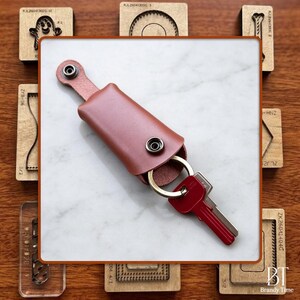 May include: A tan leather key holder with a snap button closure and a metal ring attached to a red key. The key holder features a smooth finish and a secure metal snap mechanism to keep keys organized and protected within the leather casing.