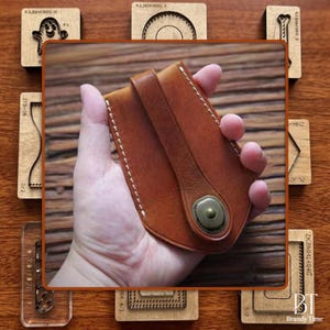 May include: A brown leather wallet with a strap and a metal button closure. The wallet is held in a hand, showcasing its design. Surrounding the wallet are wooden stamps with various shapes.