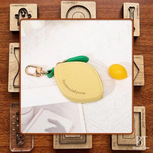 May include: A yellow lemon-shaped keychain with a green stem and gold-colored clasp. The keychain has the text "Handmade&Purse" and is next to a small lemon. The image also shows various wooden craft tools.