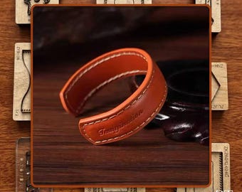 May include: A brown leather cuff bracelet with white stitching and the word "Imagination" embossed on the front. The bracelet is set against a wooden background, with various wooden craft tools surrounding it.