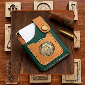May include: A green and tan leather cardholder with a brass emblem featuring an eye design. The cardholder has a snap closure and is stitched with tan thread. A white card is visible inside. The background includes wooden stamps and a pen.